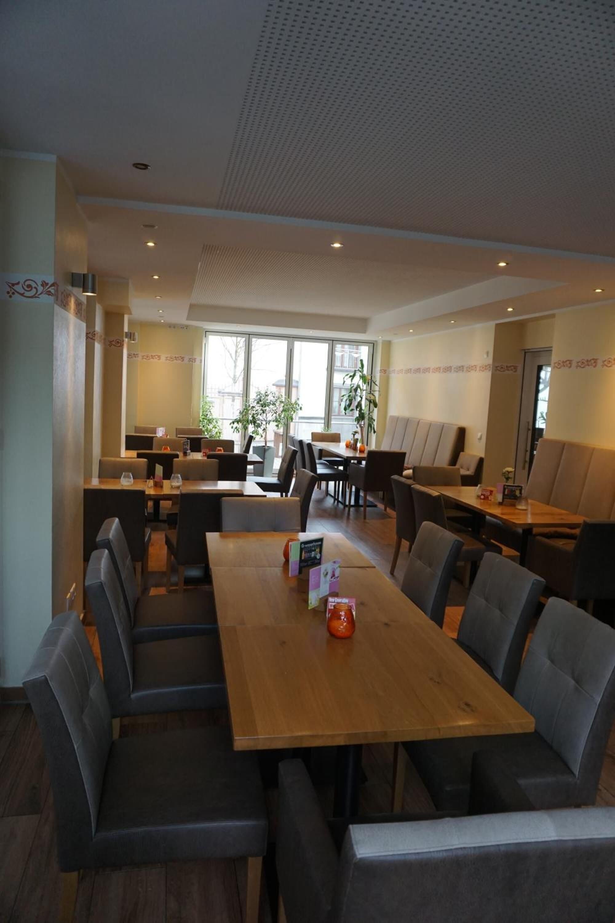 A modern restaurant with a long wooden table and cushioned chairs, surrounded by booths and a high ceiling with a perforated design.