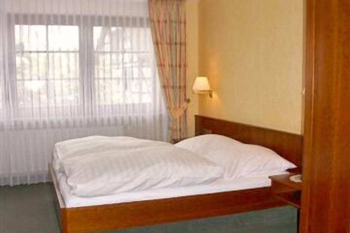 A single bed with white linens in a room with a wooden headboard, a nightstand, a lamp, and a window with sheer curtains.
