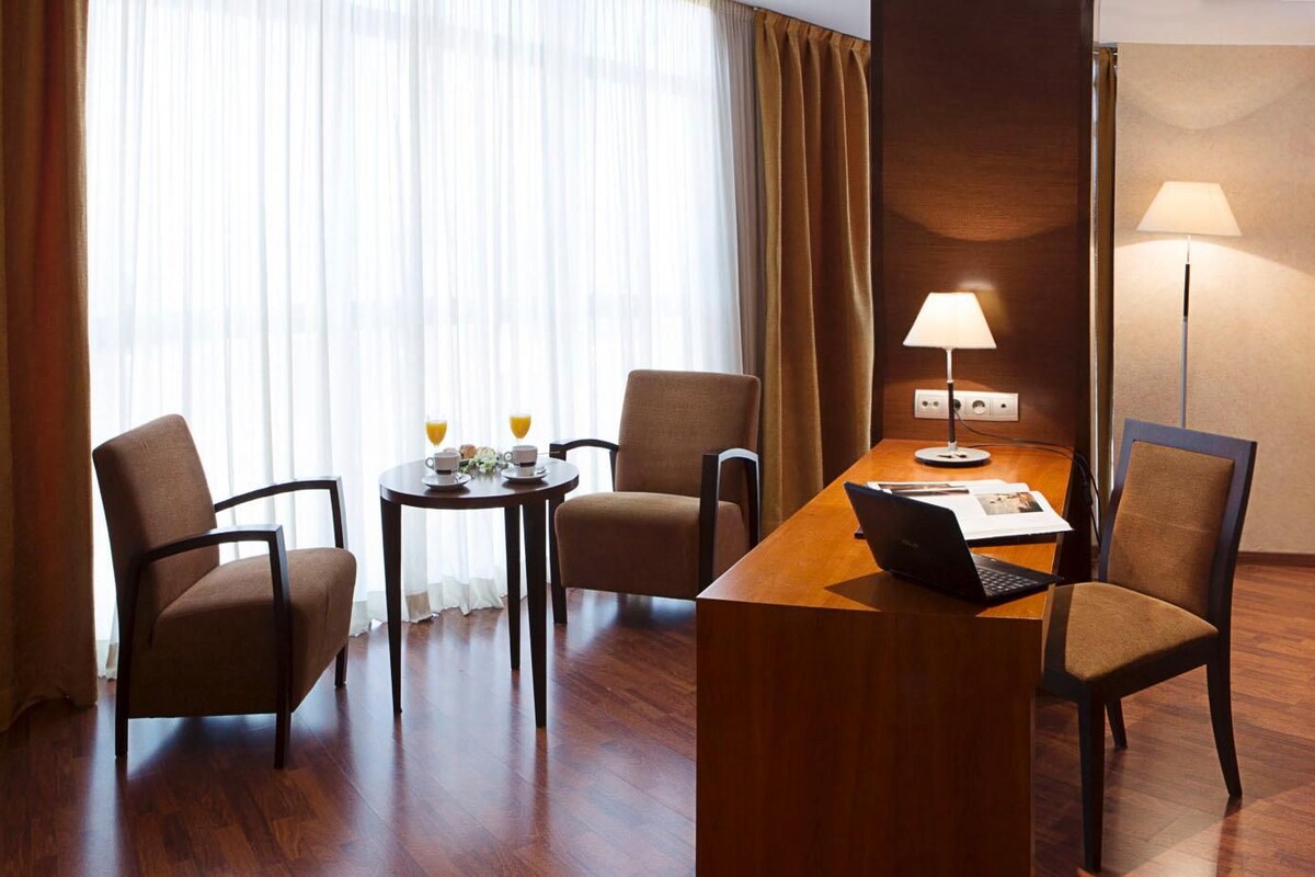 A hotel room with a bed, a desk with a laptop, two chairs, a table with breakfast items, and a lamp.