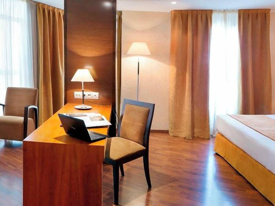 A hotel room with a bed, a desk with a laptop, a chair, and a lamp.