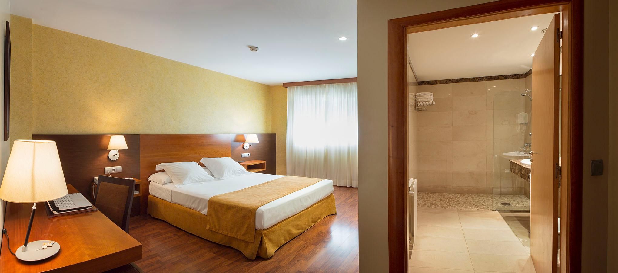 Una habitación de hotel con cama, mesitas de noche, un escritorio con lámpara y zona de ducha.