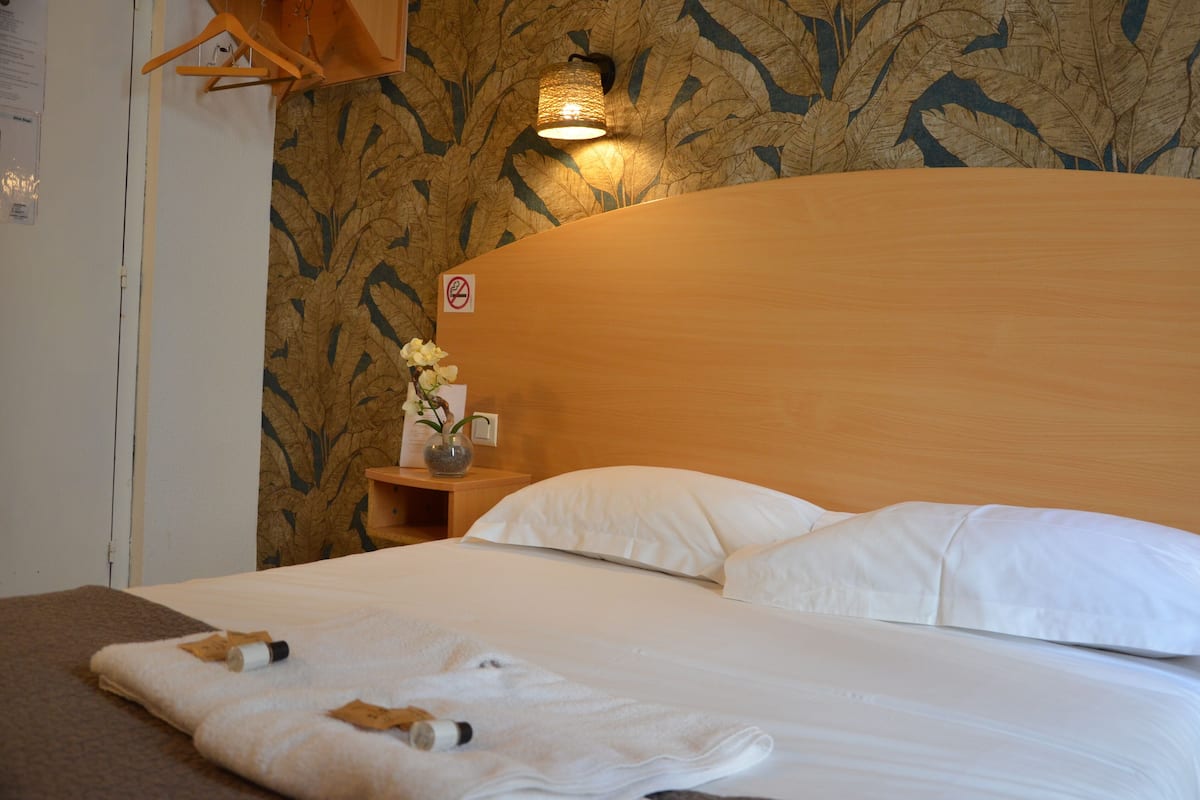Una cama bien tendida con sábanas blancas, un cabecero de madera, una mesita de noche con un florero y una pared con un estampado de hojas.