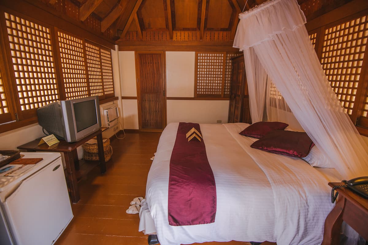A hotel room with a bed, a television on a small table, a refrigerator, and a wooden floor.