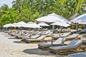 A beach with rows of sun loungers and white umbrellas, surrounded by palm trees.