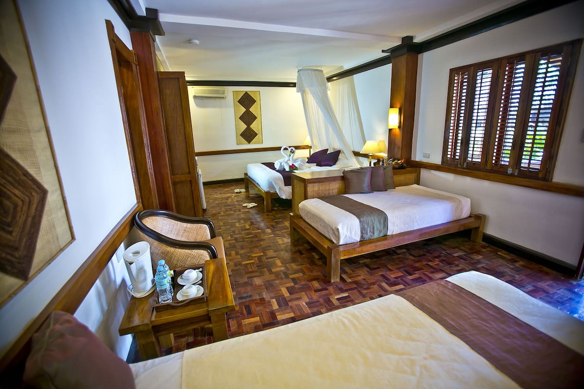A hotel room with a bed, bedside table, and a chair. There is a window with wooden shutters and a wall-mounted air conditioning unit.