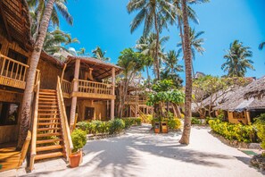 A resort with thatched-roof buildings, wooden staircases, and a sandy pathway surrounded by palm trees.