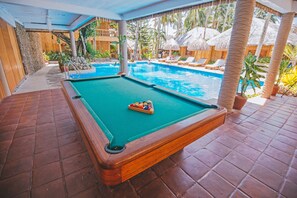 A pool table with a green felt surface, surrounded by a tiled floor and a pool area with lounge chairs and umbrellas.
