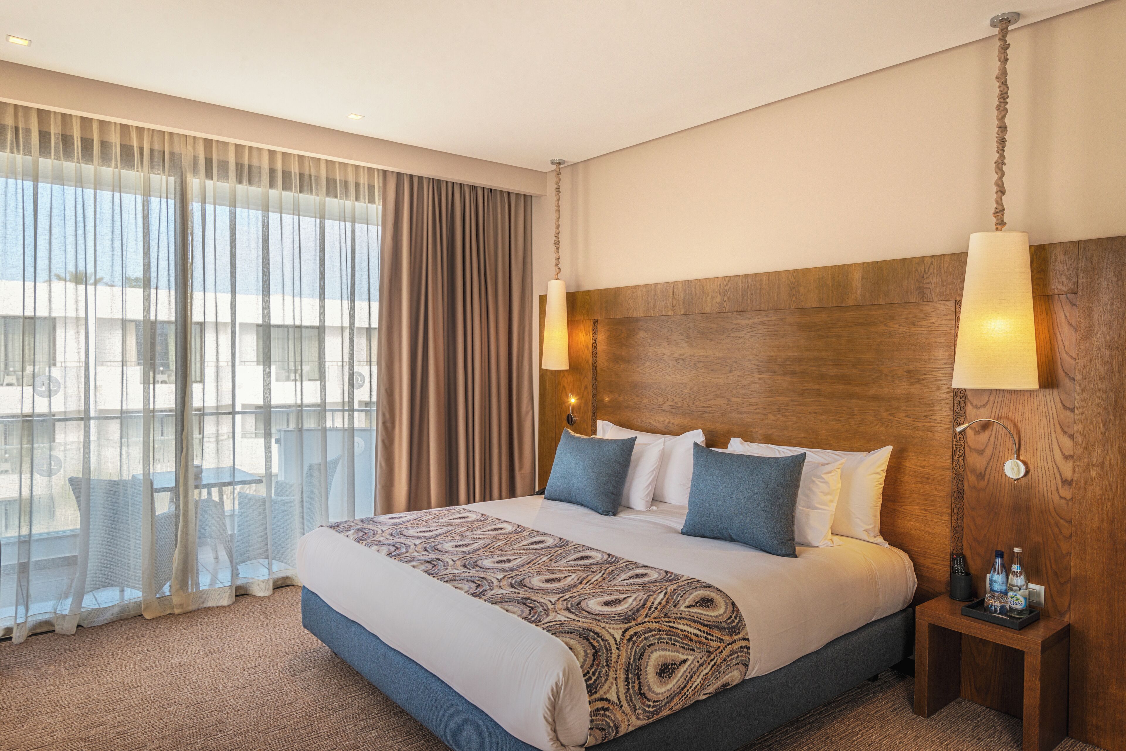 A hotel room with a large bed, a wooden headboard, two bedside lamps, a nightstand with a remote control, and a view of the outside through sheer curtains.