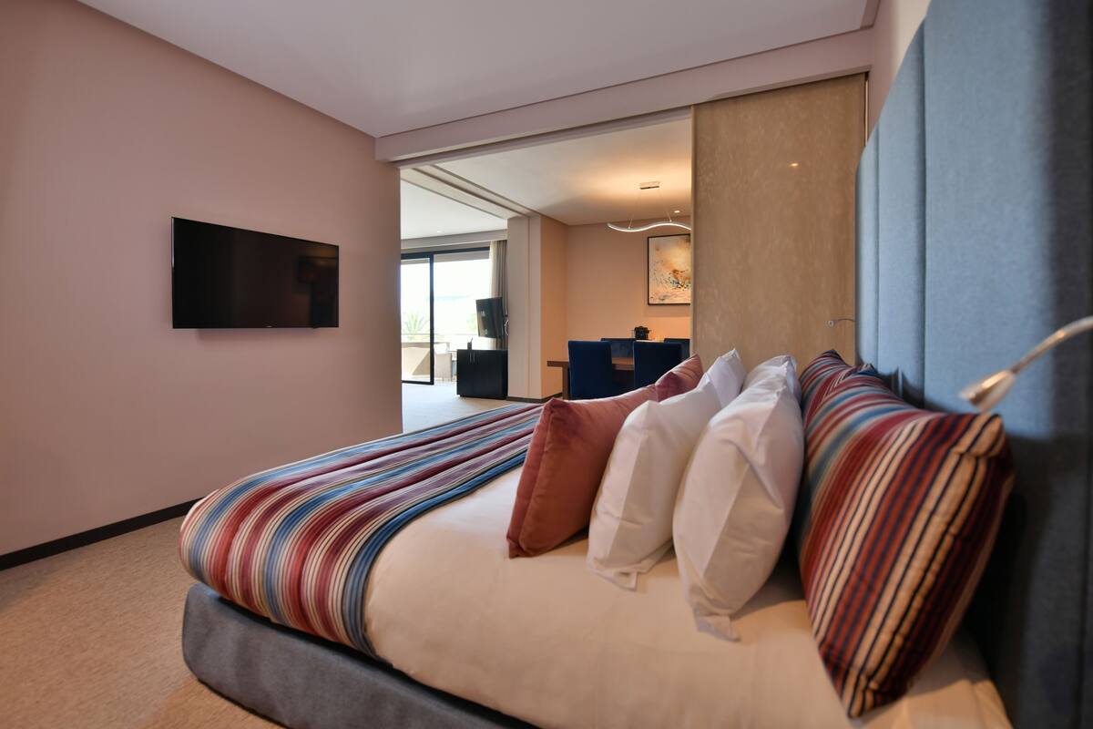 A modern hotel room with a large bed, a flat-screen TV mounted on the wall, and a view of the dining area through an open door.