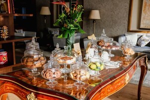 A buffet table with various desserts and pastries in glass containers, a decorative vase with flowers, and a cozy seating area with a sofa and lamps in the background.