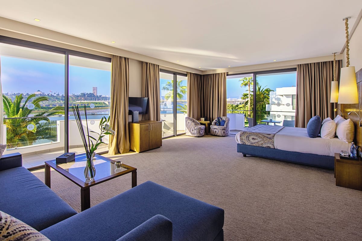 A modern hotel room with a large bed, a sofa, a TV, and a view of the city and water.