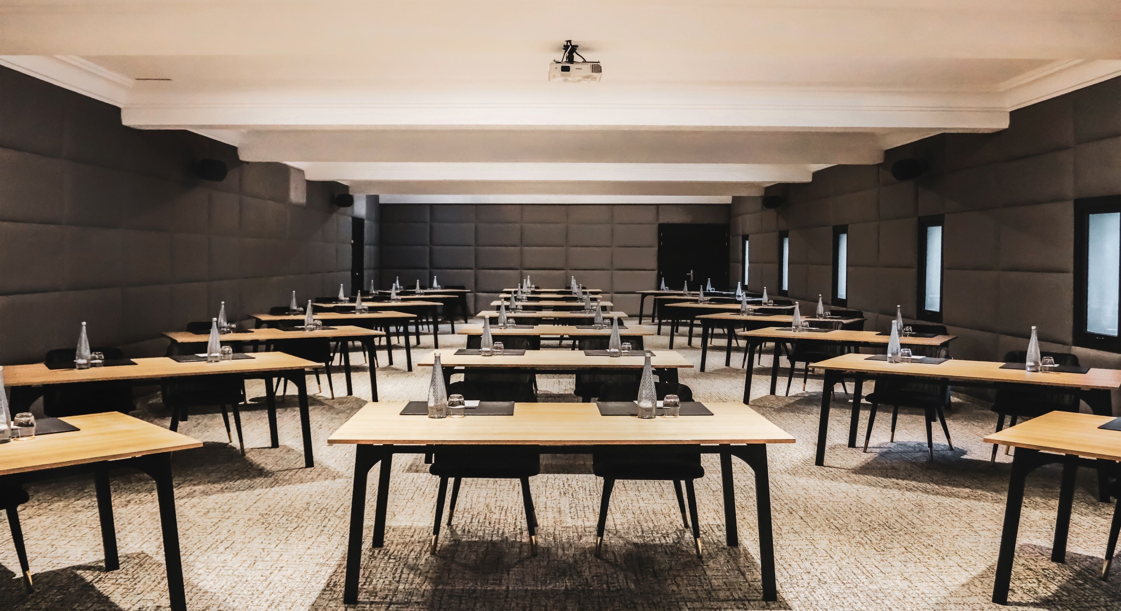 Une salle de conférence équipée de plusieurs tables et chaises, d’un projecteur au plafond et de grandes fenêtres sur le côté droit.