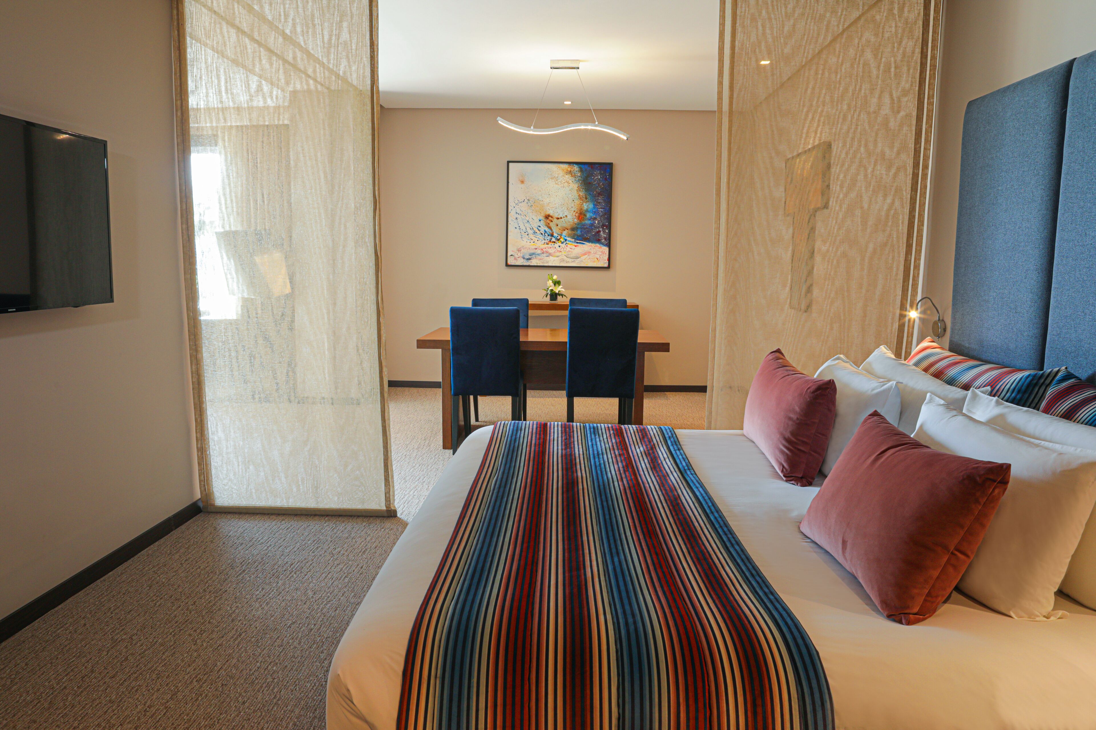A hotel room with a large bed, a dining table with chairs, and a TV mounted on the wall.