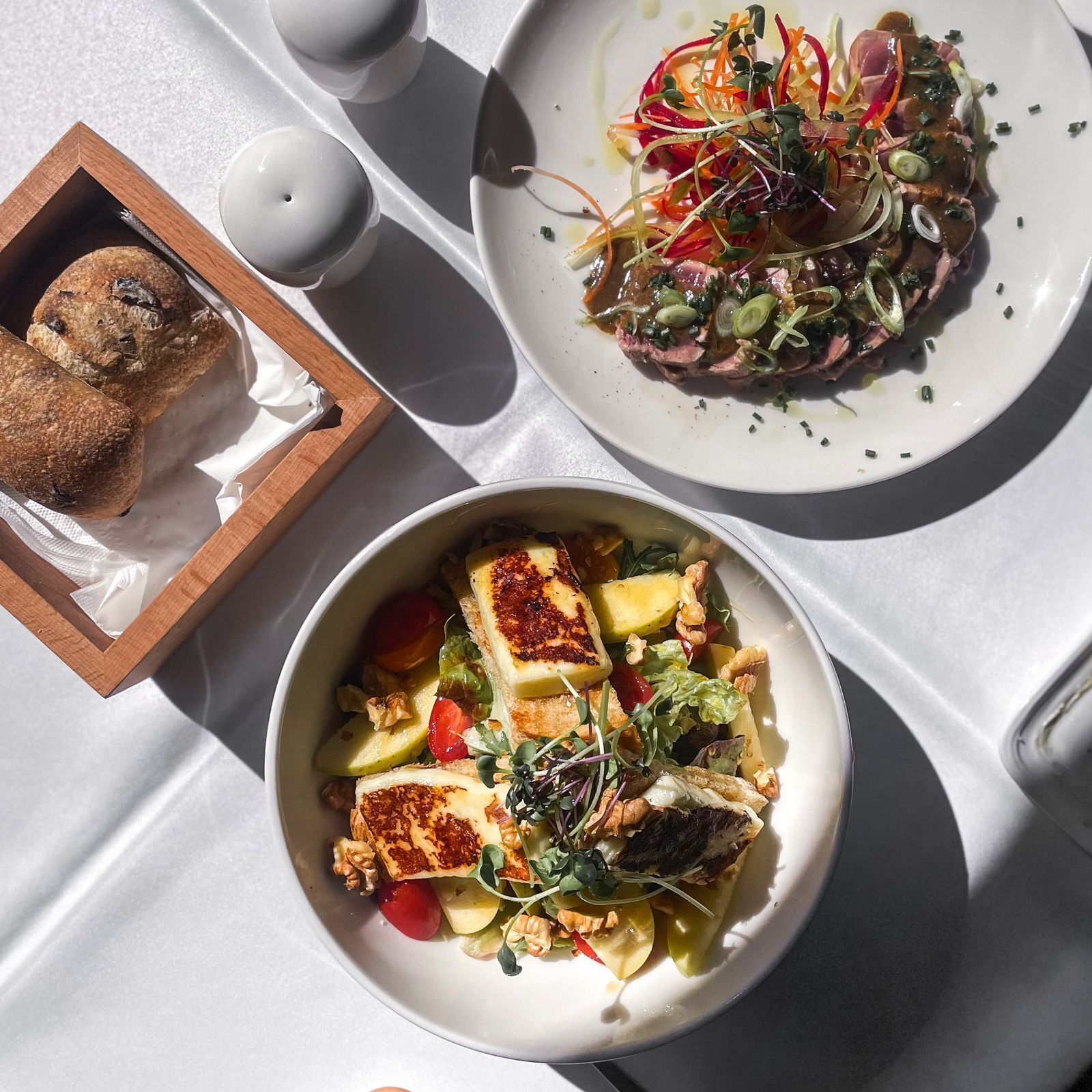 A table with three plates of food, a wooden box with bread, and salt and pepper shakers.