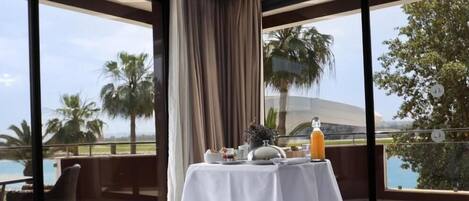Una habitación de hotel moderna con una cama grande, una mesa de comedor con alimentos para el desayuno y vistas a una piscina y palmeras.