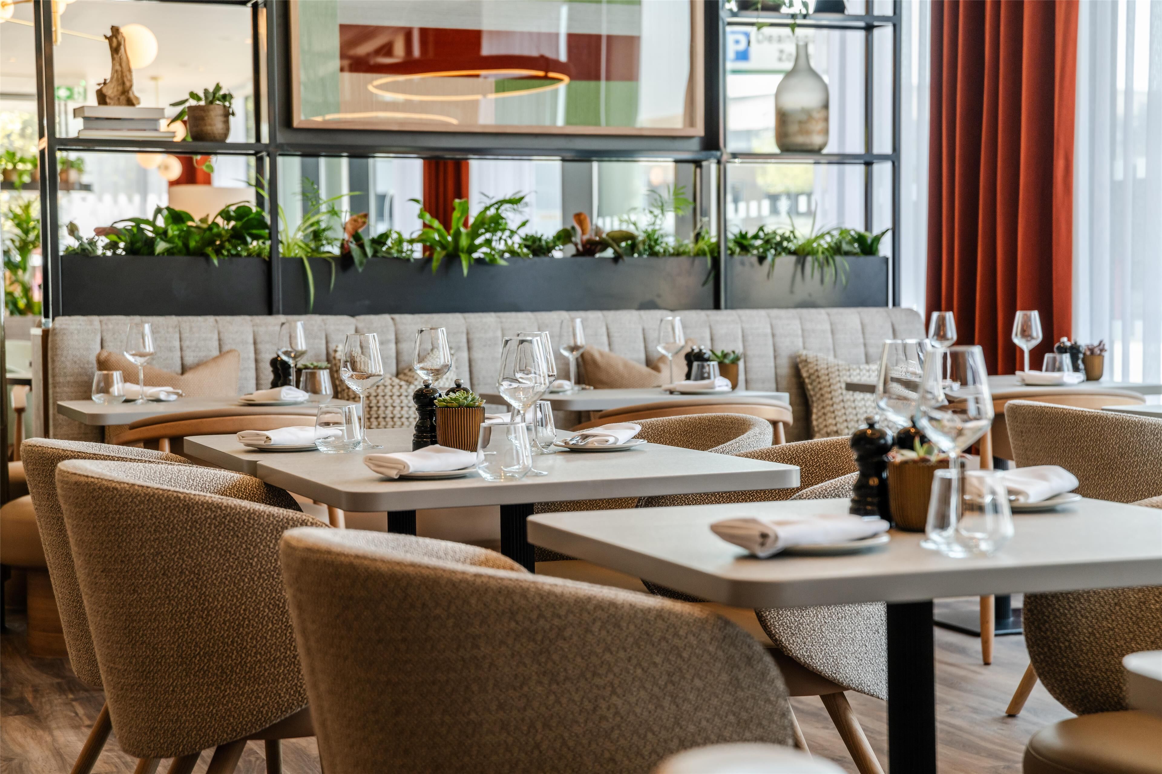 Un restaurante con una larga zona de asientos tipo banco, varias mesas y una diversidad de plantas al fondo.