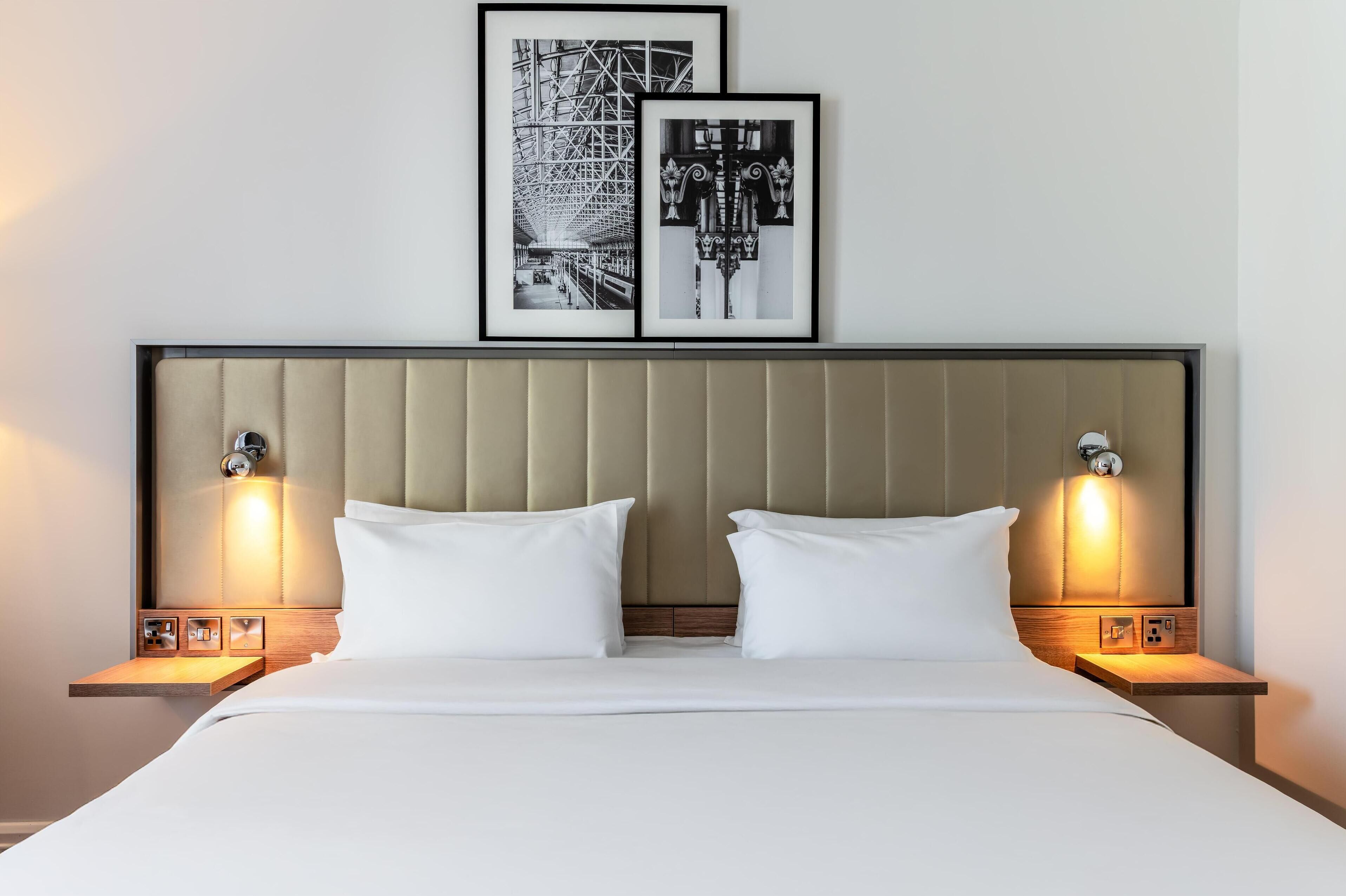Una cama bien hecha con sábanas blancas, dos mesitas de noche con lámparas, y dos fotografías en blanco y negro enmarcadas en la pared sobre la cama.