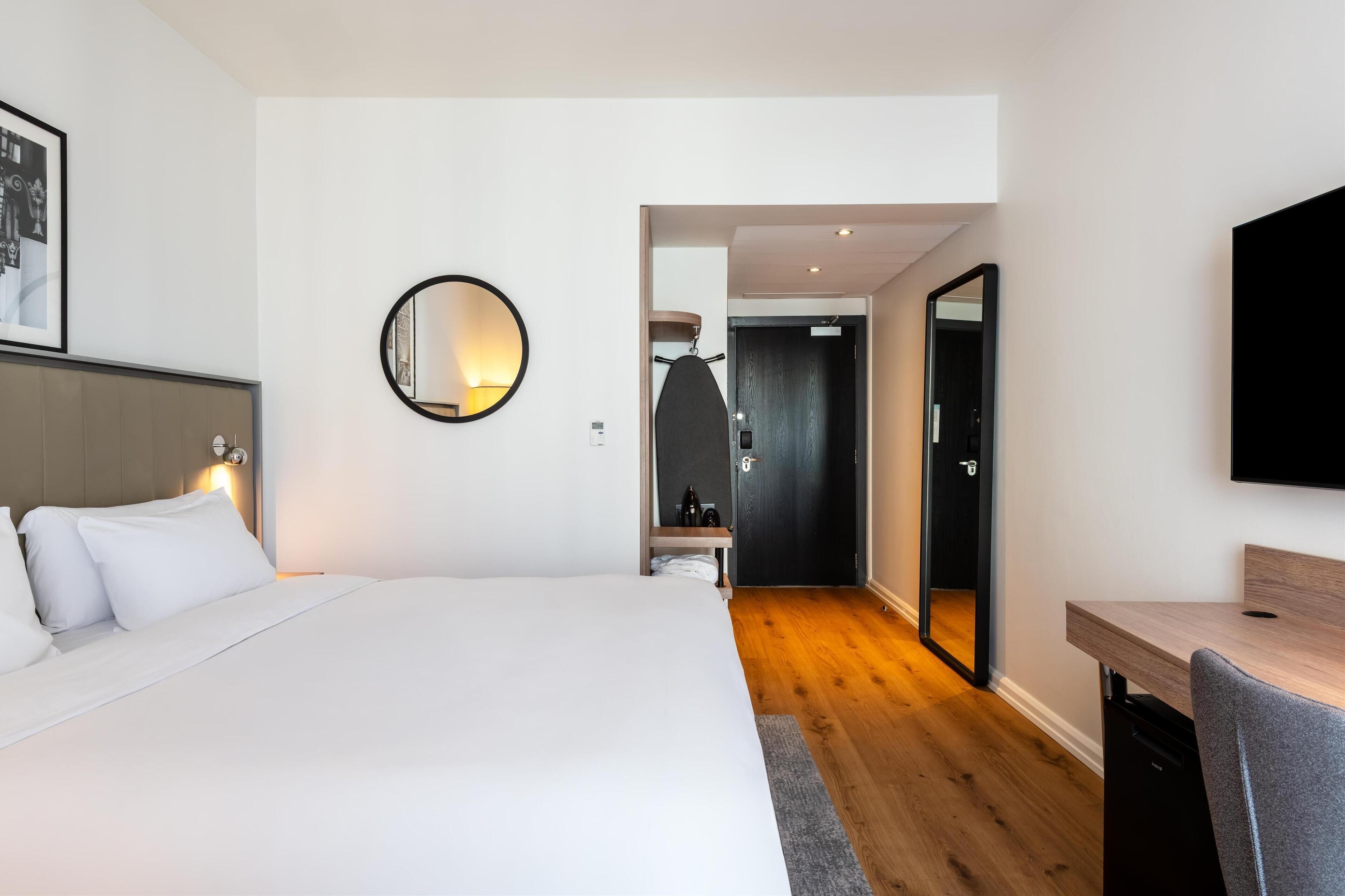 A modern hotel room with a large bed, a round mirror, a desk, and a wardrobe.
