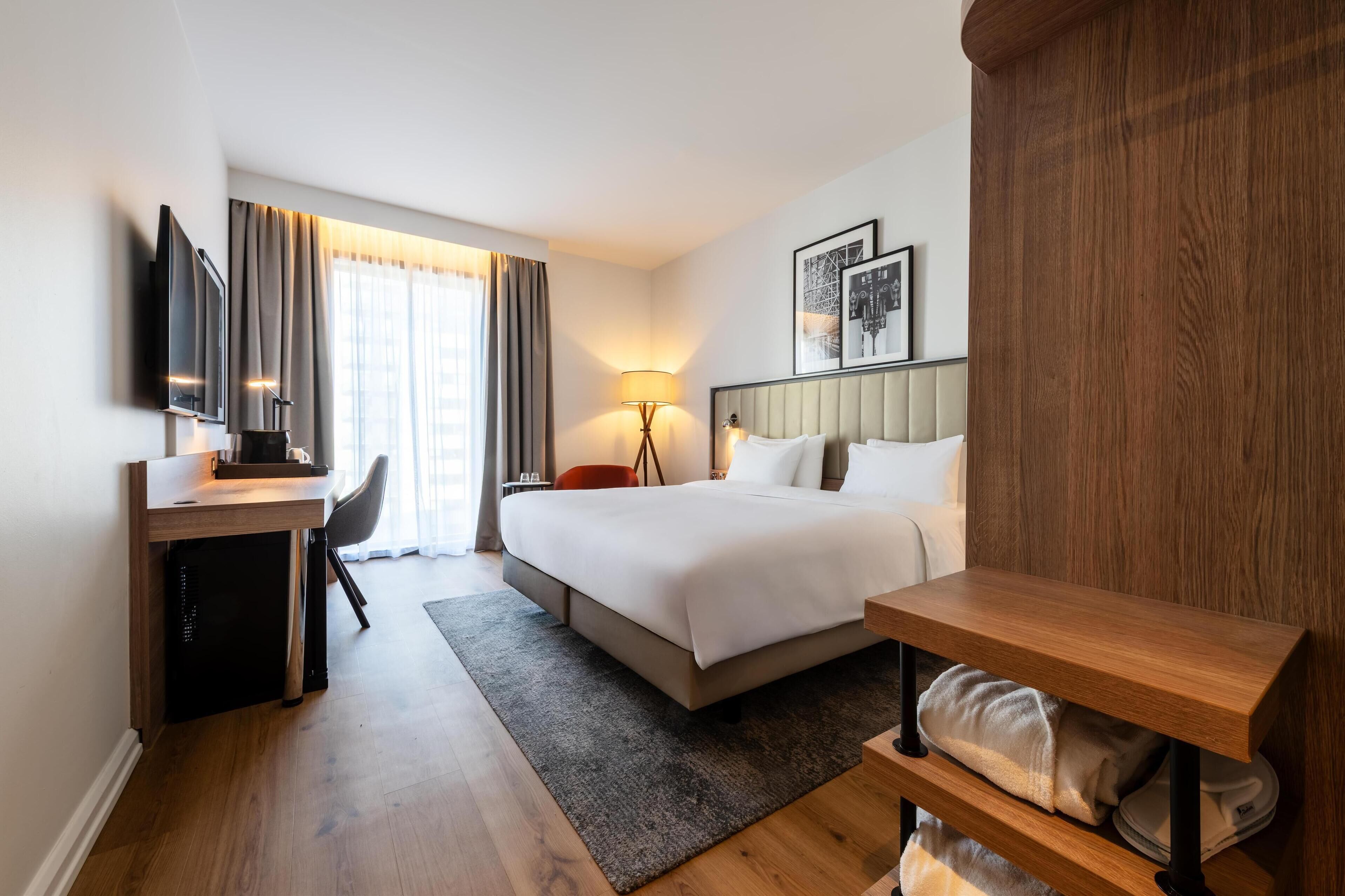 A hotel room with a bed, a desk, a chair, a lamp, and a TV.