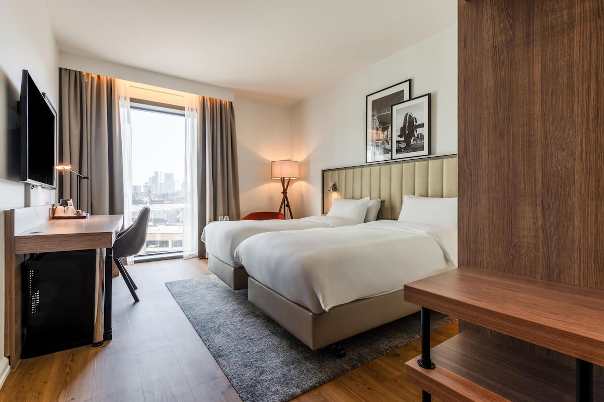 A hotel room with a bed, a desk, a chair, a TV, a lamp, and a city view.