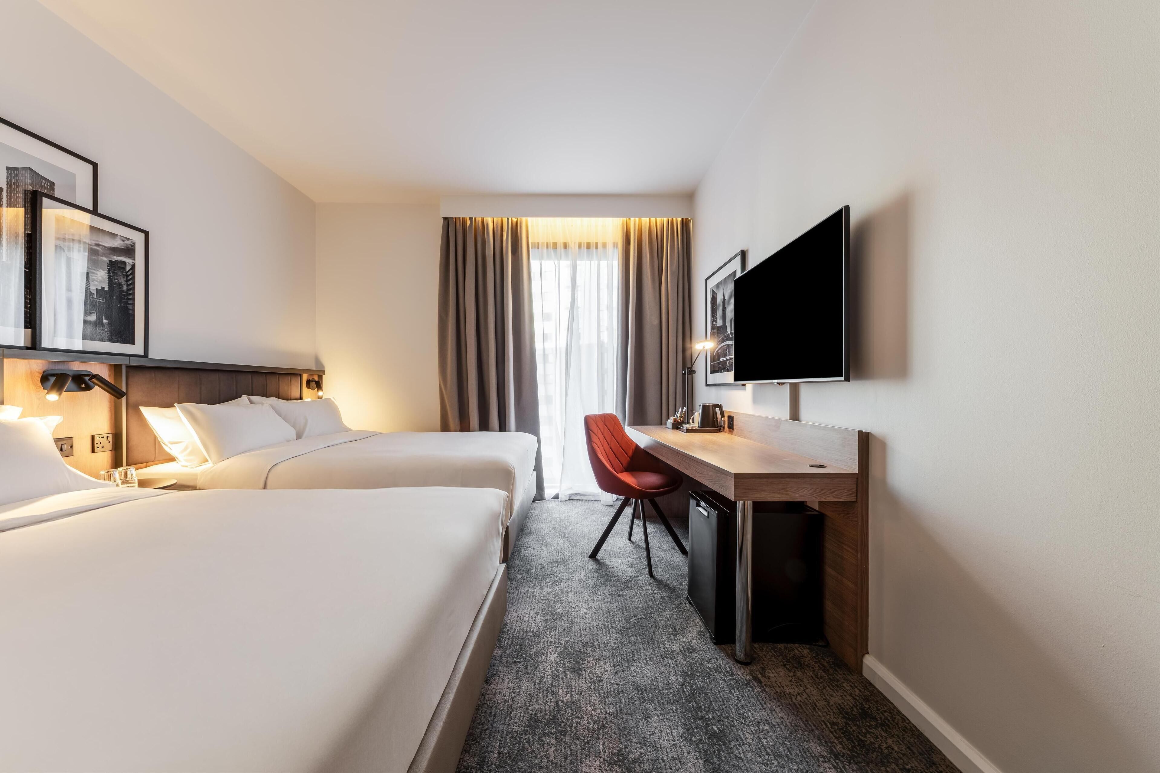 A hotel room with two beds, a desk, a chair, a TV, and a window with curtains.