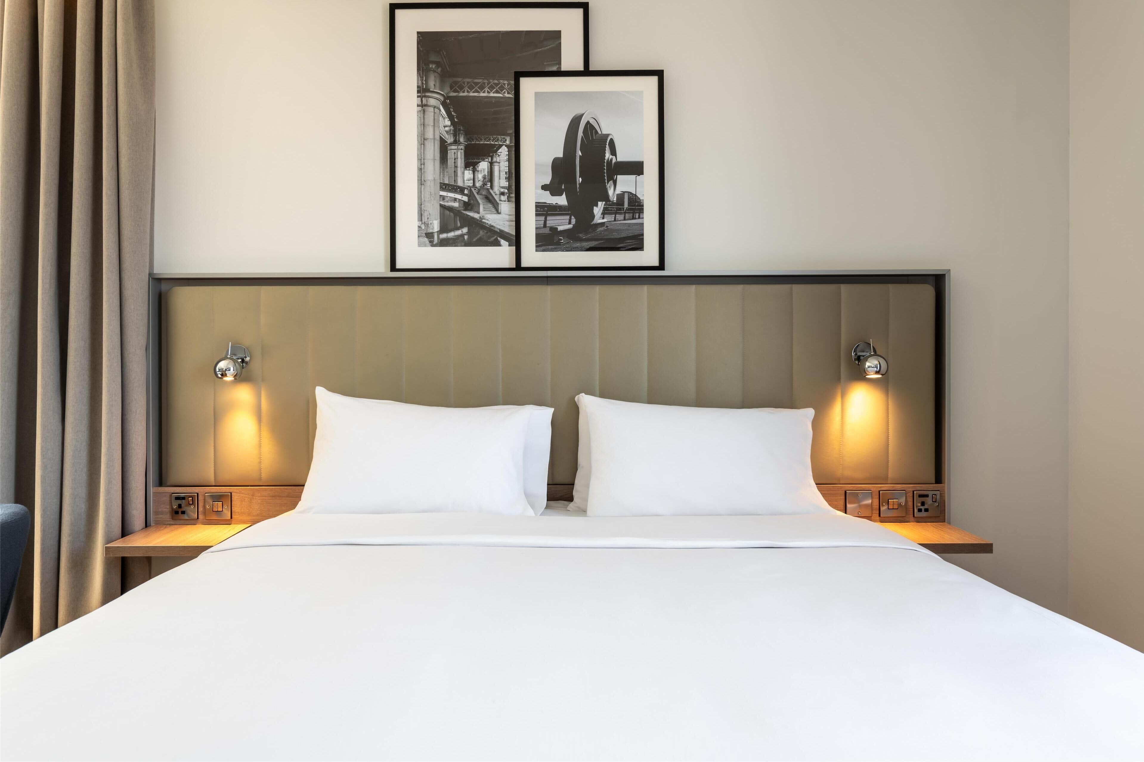 A hotel room with a large bed, two bedside tables with lamps, a headboard, and two framed pictures on the wall.
