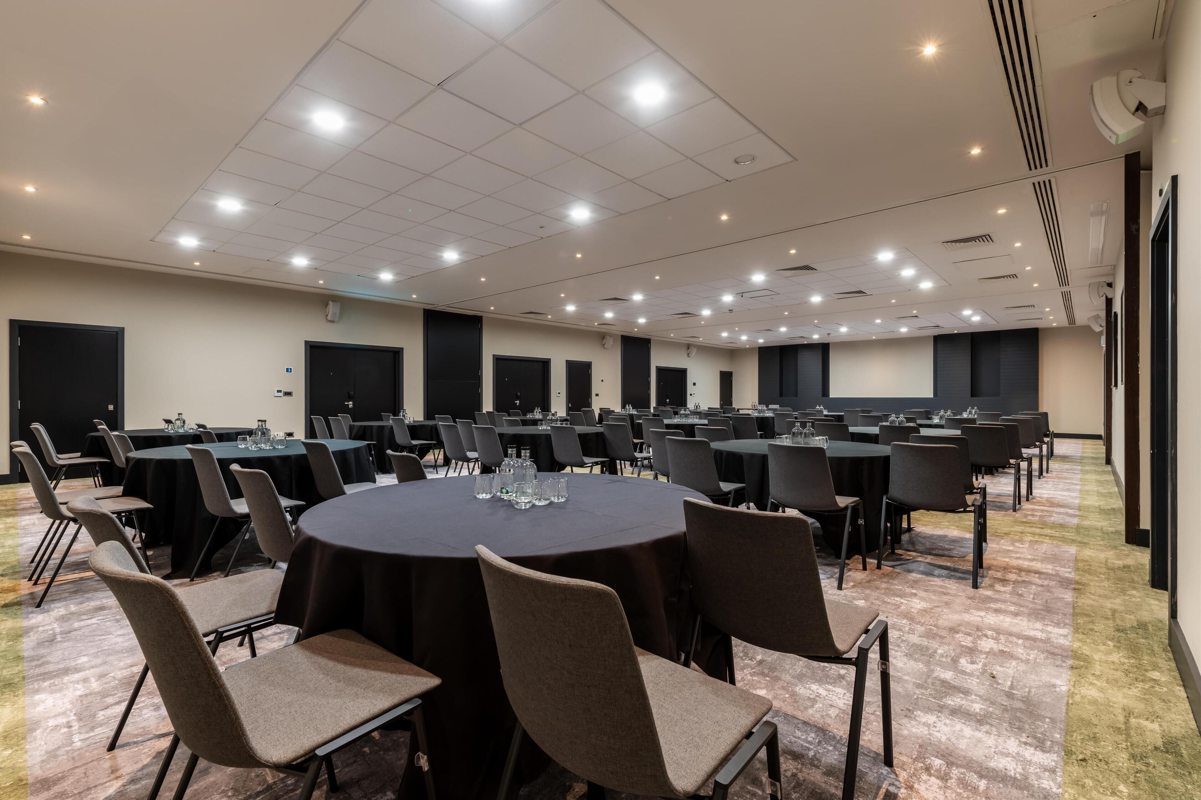 Un salón de conferencias con varias mesas y sillas, equipado con vasos de agua y una pantalla para proyector.