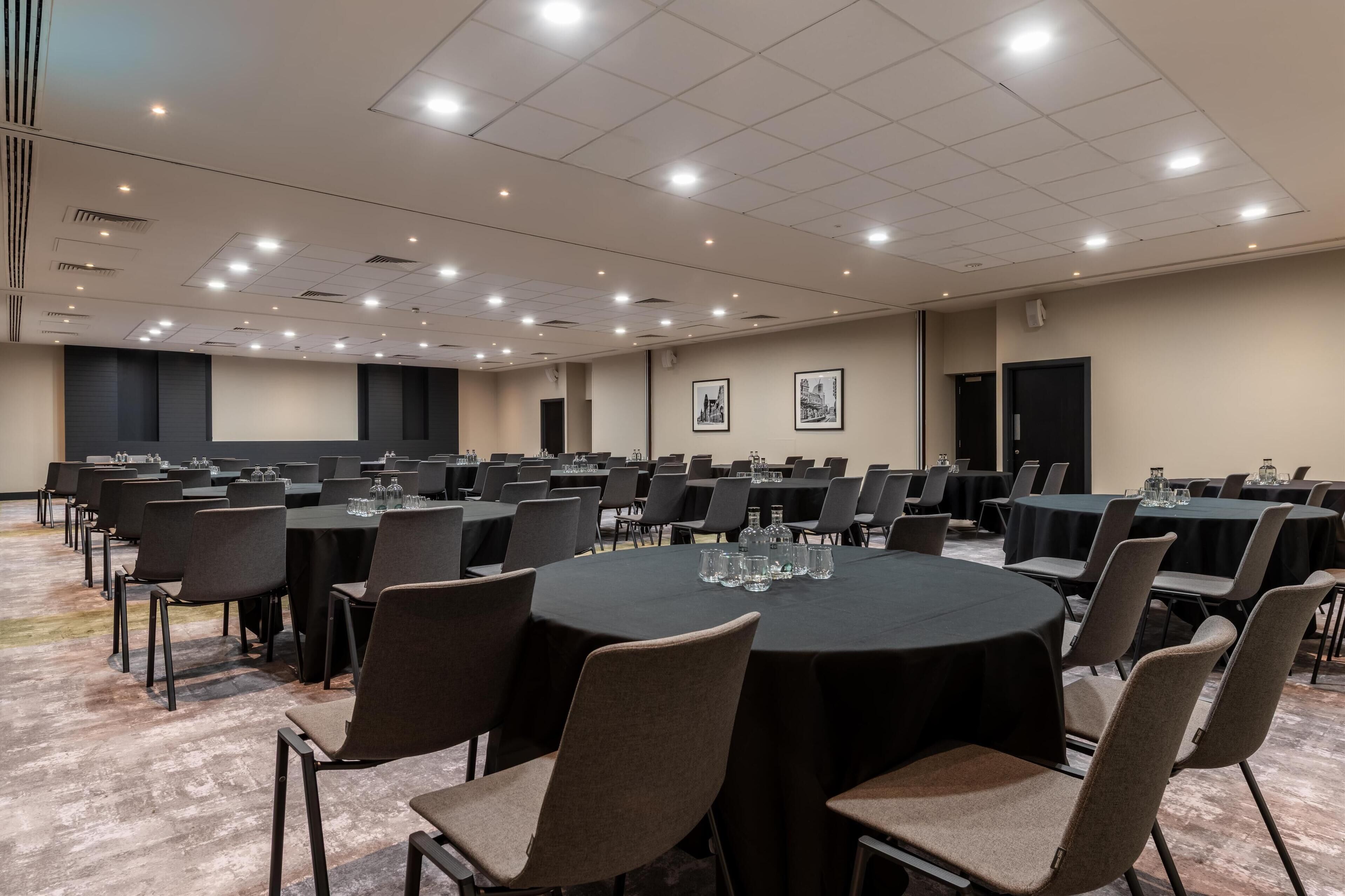 Un salón de conferencias con varias mesas y sillas, equipado con vasos de agua y una pantalla para proyector.