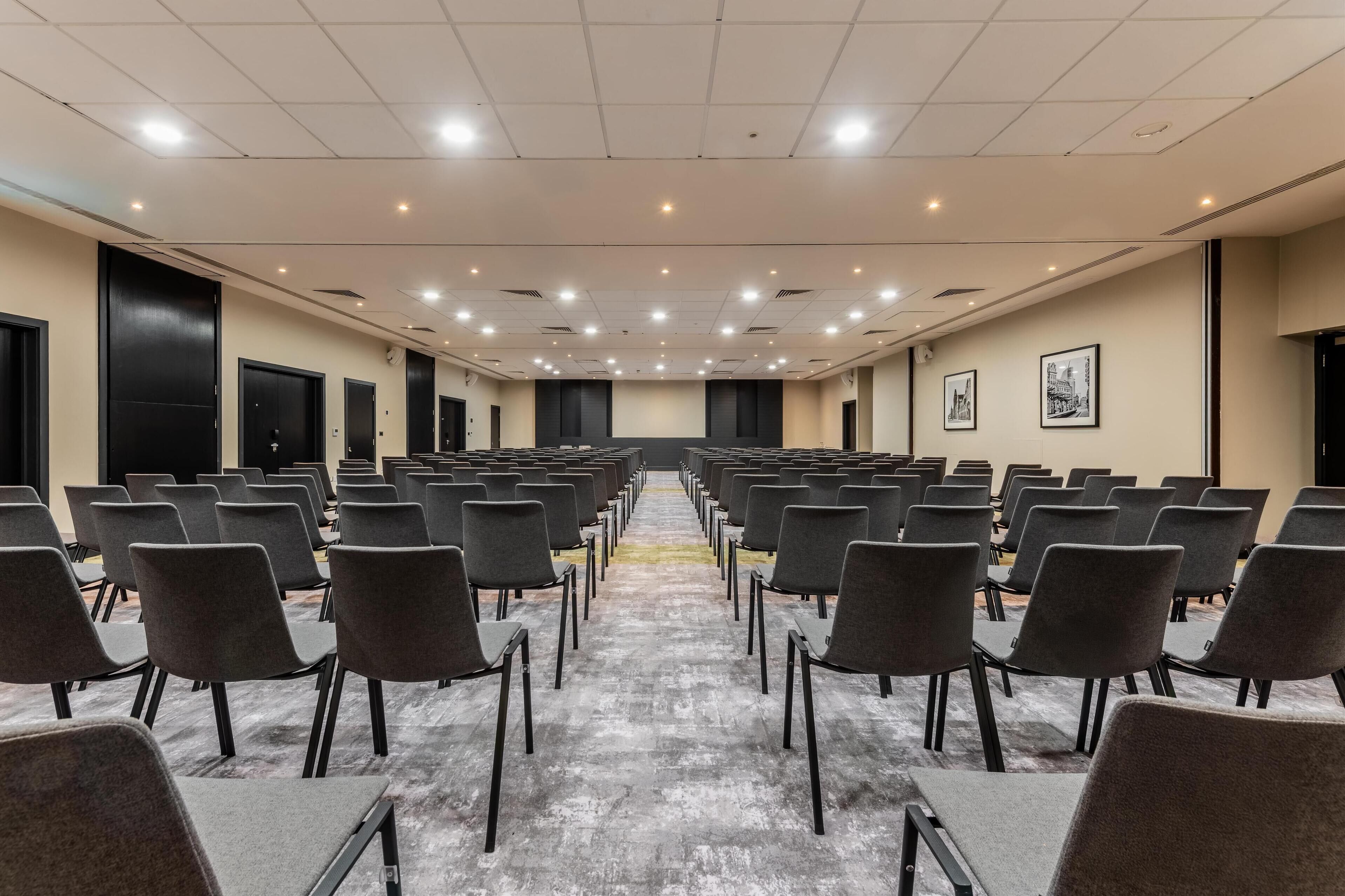 A conference room with rows of chairs, a stage, and a carpeted floor.