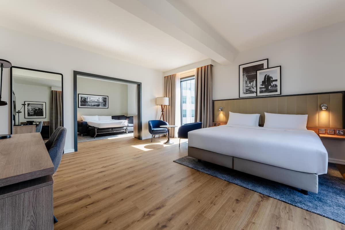 A hotel room with a large bed, a desk, a chair, a small table, and a sofa. There is a window with curtains, a lamp, and framed pictures on the wall.