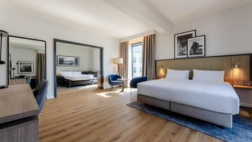 A hotel room with a large bed, a desk, a chair, a small table, and a sofa. There is a window with curtains, a lamp, and framed pictures on the wall.