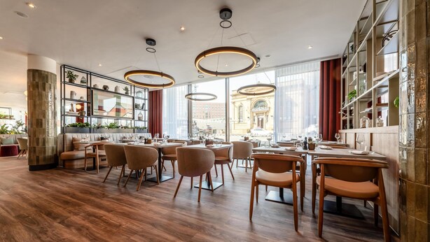 En moderne restaurant med trægulve, runde borde og stole. Store vinduer med udsigt til en bygning.