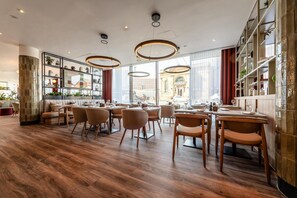 A modern restaurant with wooden floors, round tables, and chairs. There are large windows with a view of a building outside. - Radisson Hotel Manchester City Centre, A Verified Net Zero Hotel (Manchester)