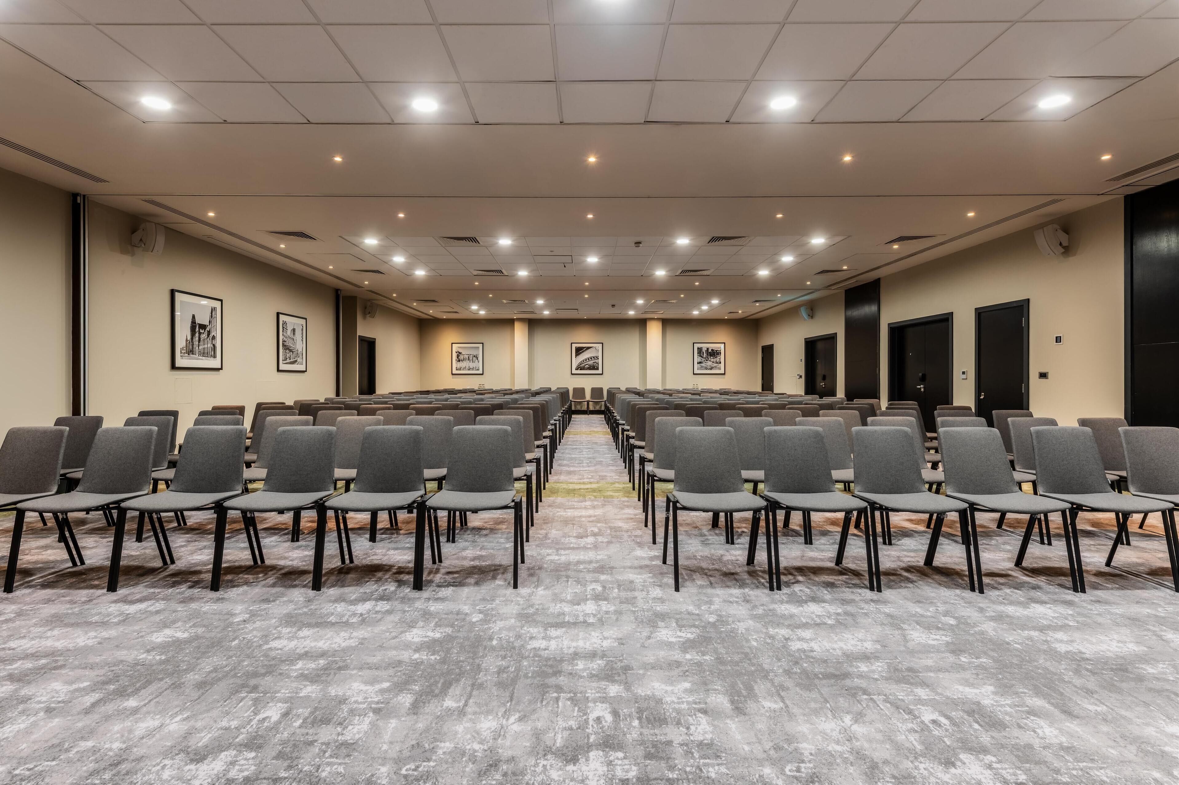 Uma sala de conferências com fileiras de cadeiras, piso coberto por carpete e quadros nas paredes.