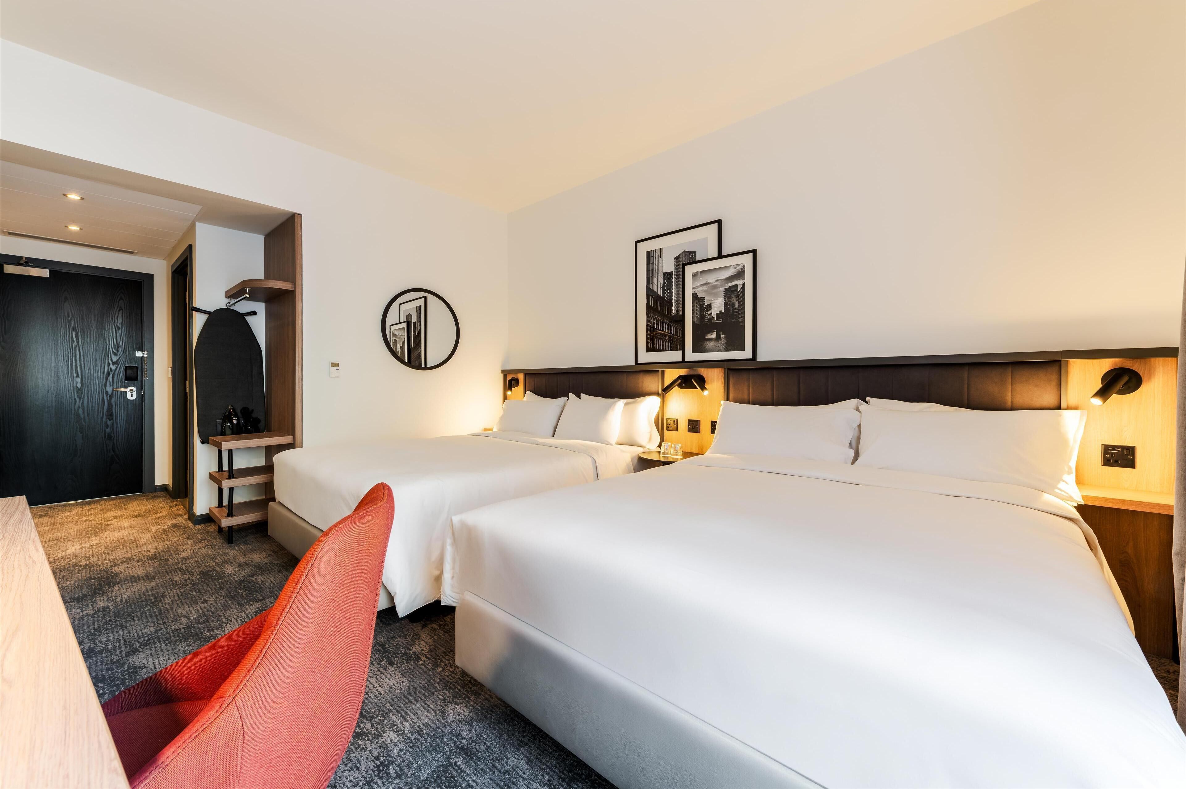 A hotel room with two beds, a desk, a chair, and a coat rack.