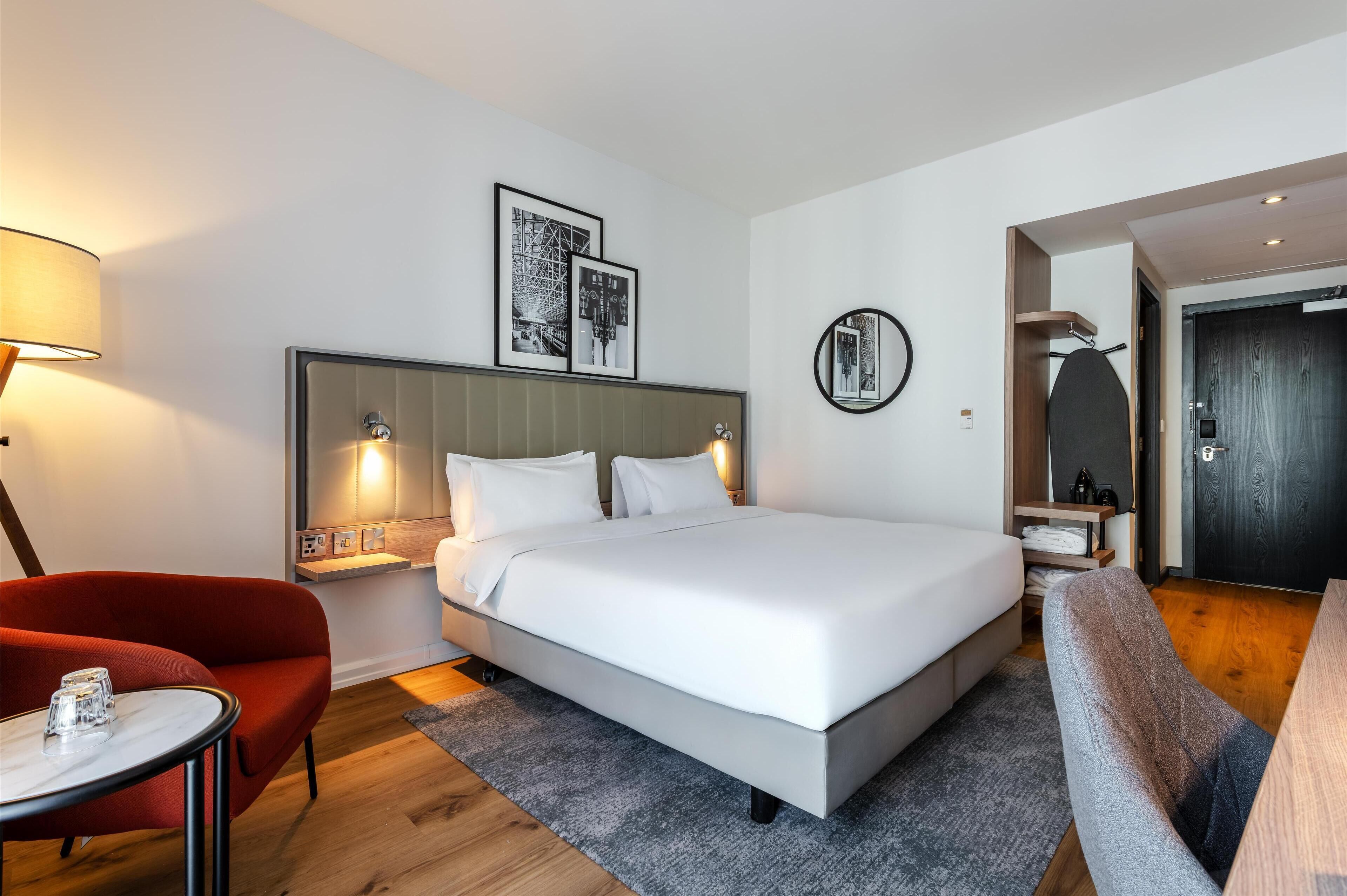 A modern hotel room with a large bed, a red armchair, a small table, and a round mirror.