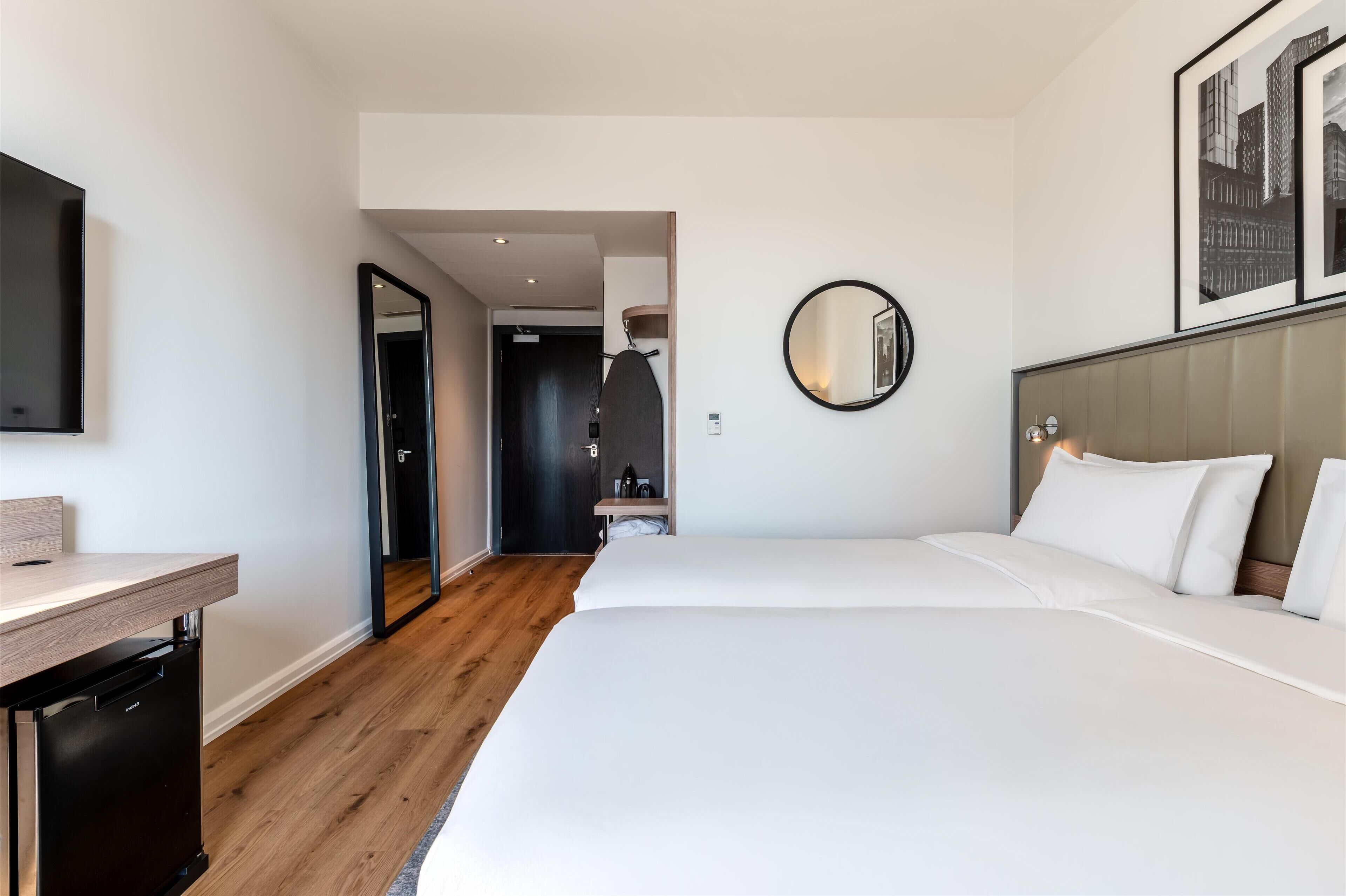 A hotel room with a bed, a desk, a mirror, and a door leading to another room.