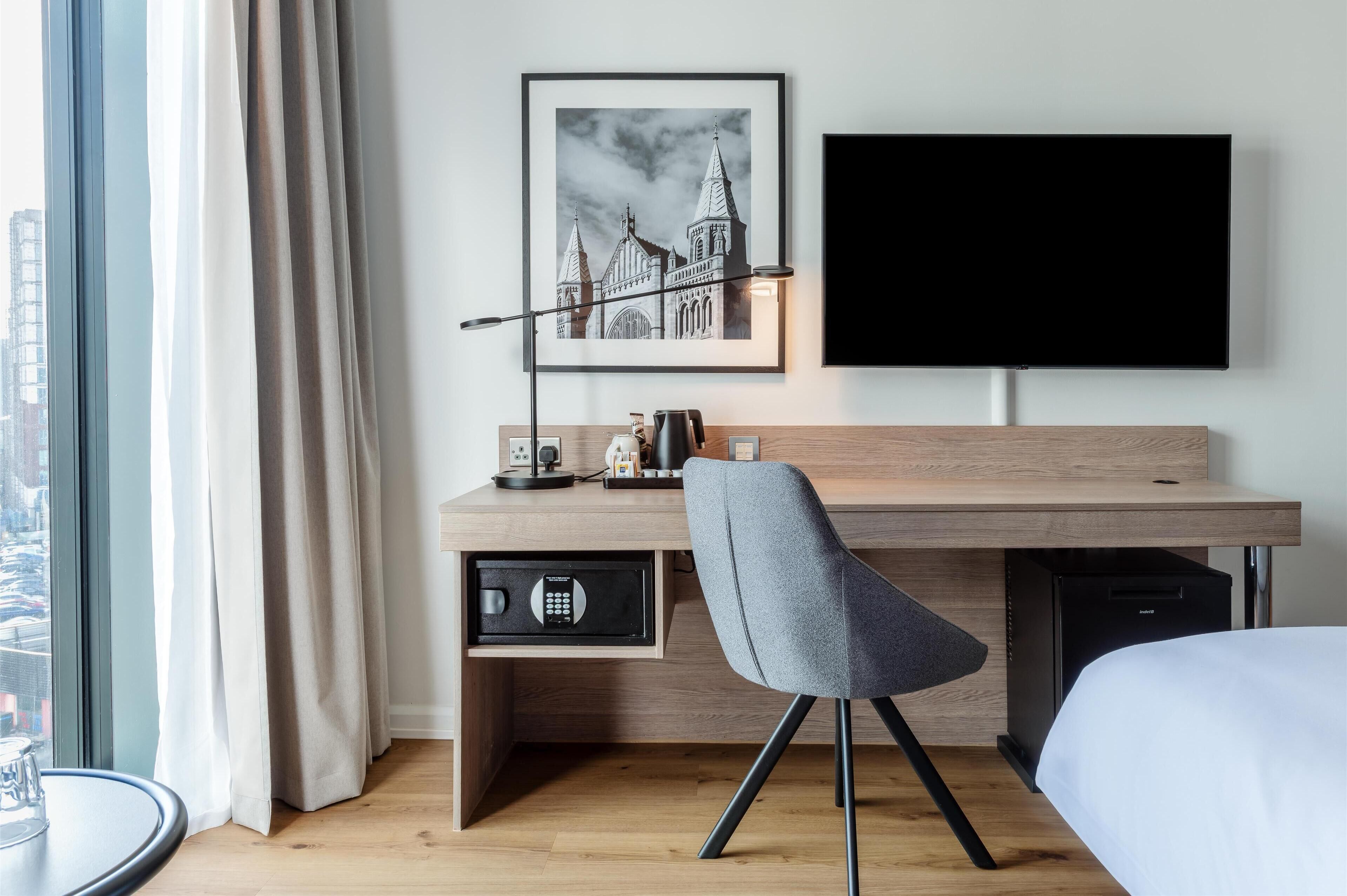 Habitación de hotel moderna con un escritorio de madera, una silla gris, una caja fuerte negra, un televisor de pantalla plana y un cuadro con una imagen de un edificio.