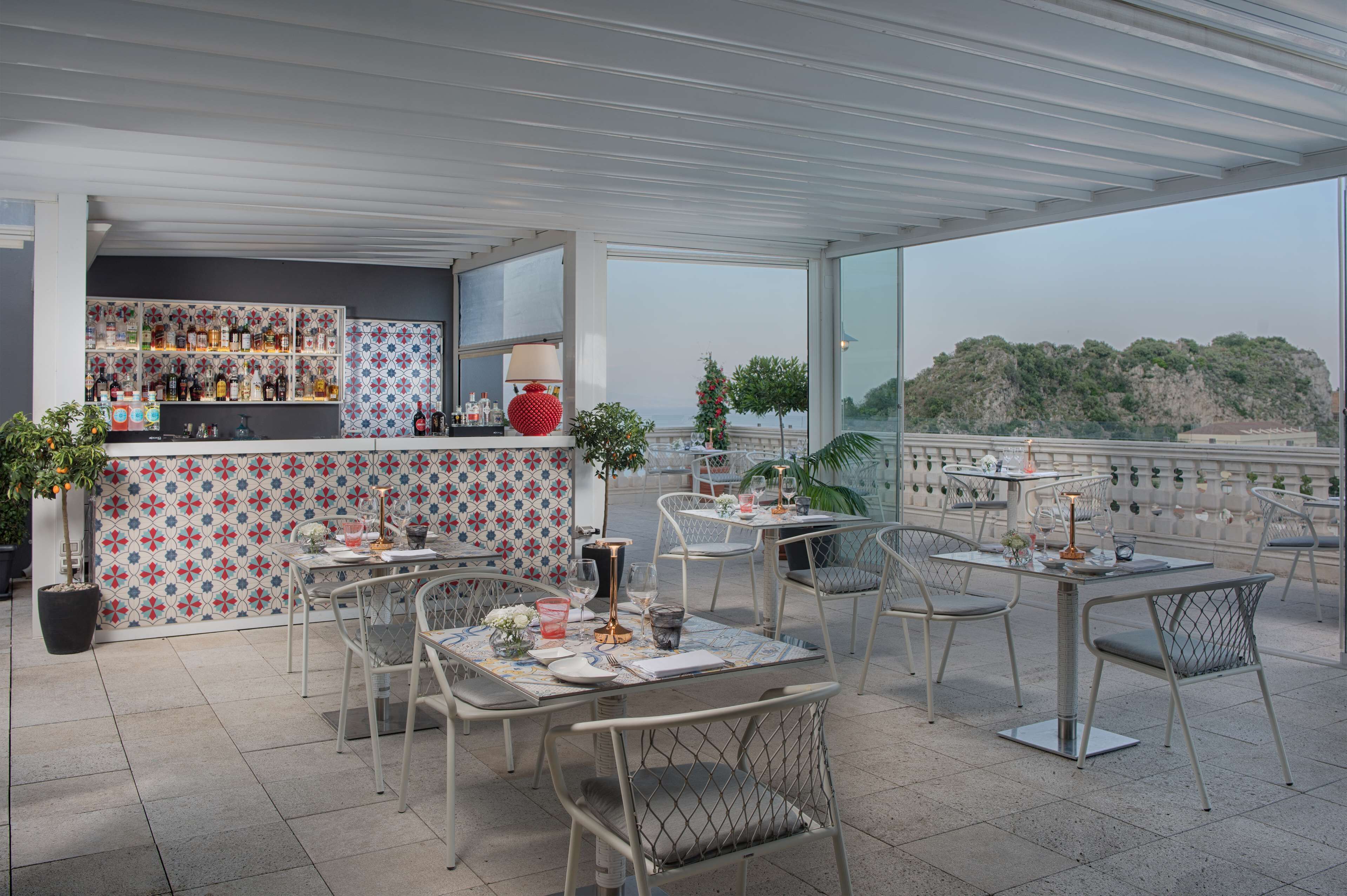 Een dakterras met betegelde vloer, metalen stoelen en tafels gedekt voor diner. Een bar met verschillende flessen en een rode decoratie is zichtbaar.