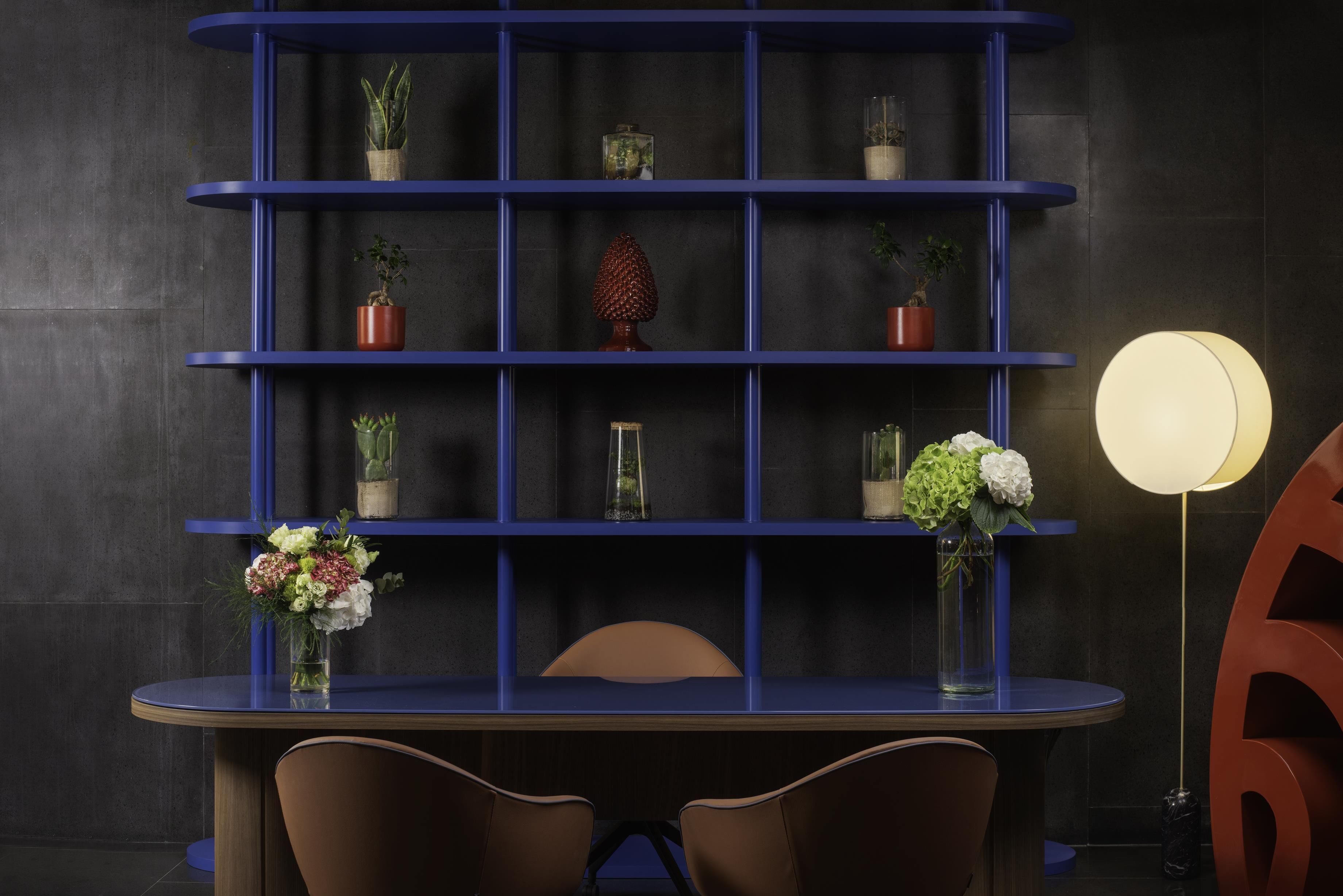 Ein modernes Interieur mit einer blauen Regaleinheit, die verschiedene Pflanzen und Blumen zeigt, einem runden Tisch mit Stühlen und einer großen dekorativen Zahl.