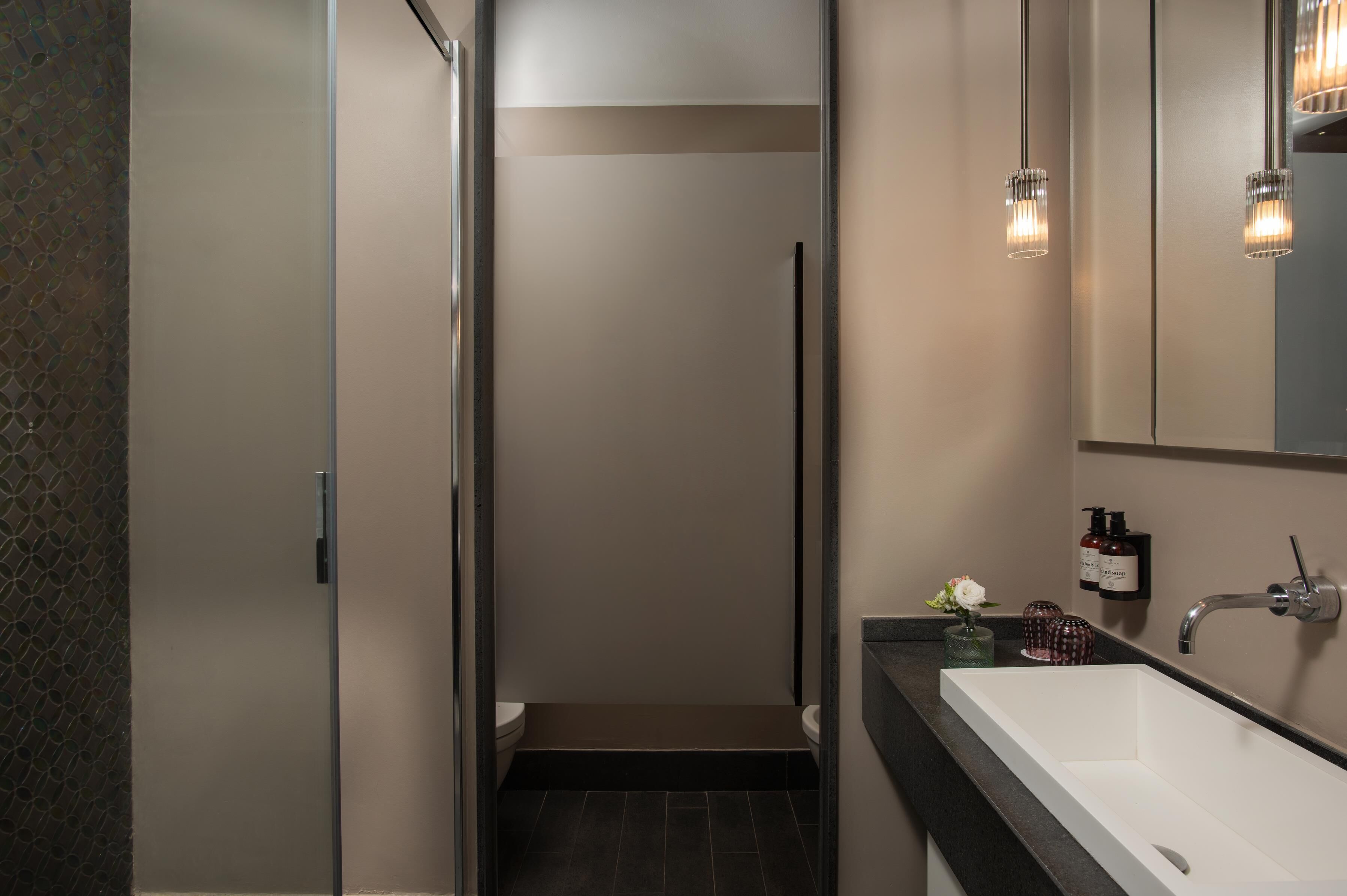 A modern bathroom with a white sink, dark tiles, a glass shower enclosure, and a mirror with pendant lights.