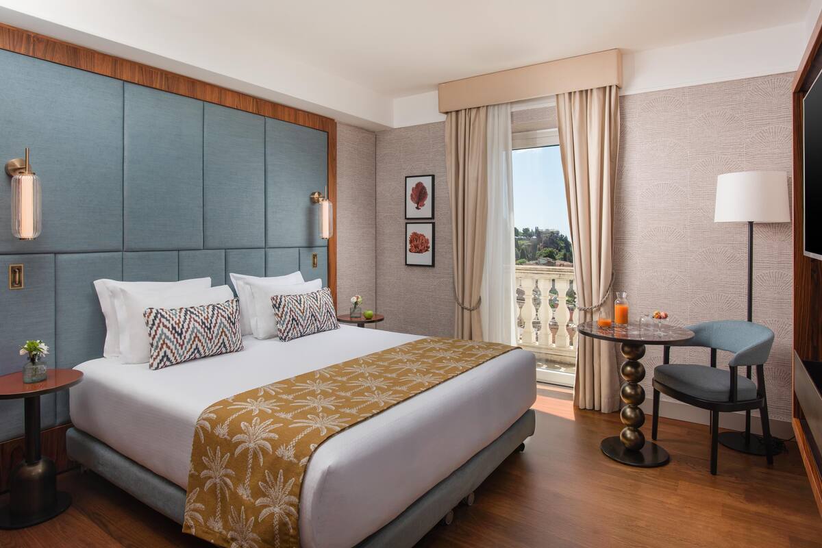 A hotel room with a large bed, a small table with a chair, a TV, and a balcony with curtains.