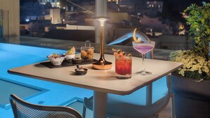 Una mesa con una bebida, una lámpara y un cuenco de fruta en un balcón con vistas a un paisaje urbano nocturno.