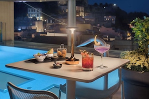 A table with a drink, a lamp, and a bowl of fruit on a balcony overlooking a cityscape at night. - NH Collection Taormina (Taormina)