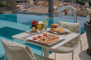Una mesa de comedor con comida, una bebida y vista a la piscina.