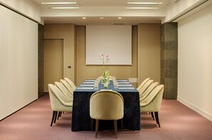 Un salón de conferencias con una larga mesa, seis sillas, una pantalla de proyector y un arreglo floral.