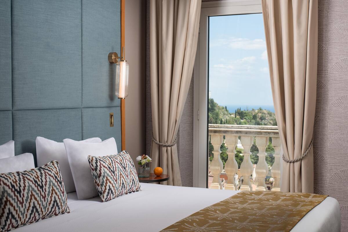 A hotel room with a large bed, a view of the sea, and a balcony with a railing.