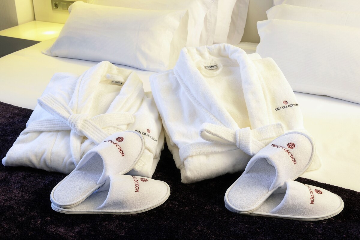 A neatly made bed with white bedding and pillows, a pair of white slippers with a logo, and a folded white robe.