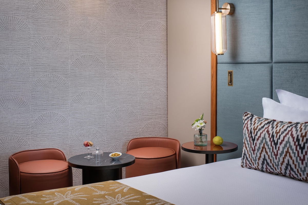 Una habitación de hotel con una cama, dos sillas, una mesita con un frutero, un jarrón con flores y una pared con textura.