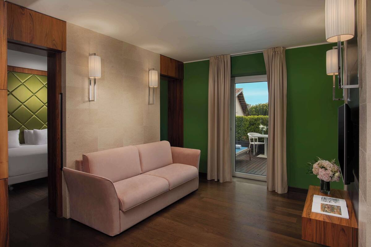 A modern hotel room with a pink sofa, wooden accents, a large window with a view of a deck and greenery, and a small table with a vase of flowers.